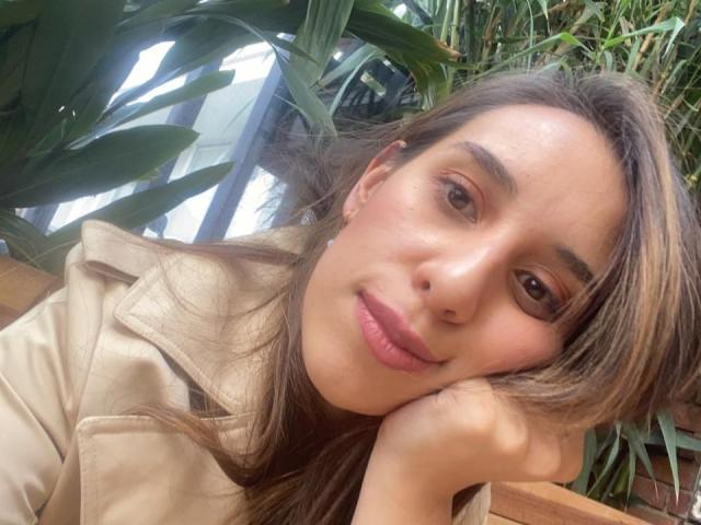 BellaBrownX with pouty lips, brown eyes, and light brown hair, head resting on hand, wearing a beige coat, surrounded by green plants.