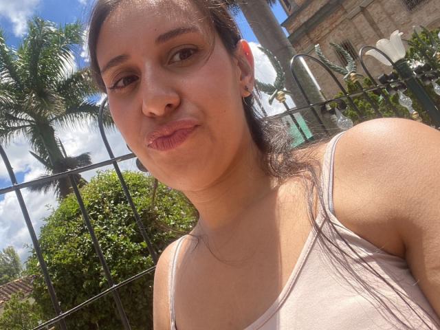 BellaBrownX with puffy lips, light brown skin, brown eyes, and small tits, wearing a pink tank top, standing outdoors, sunny day, black fence in background.