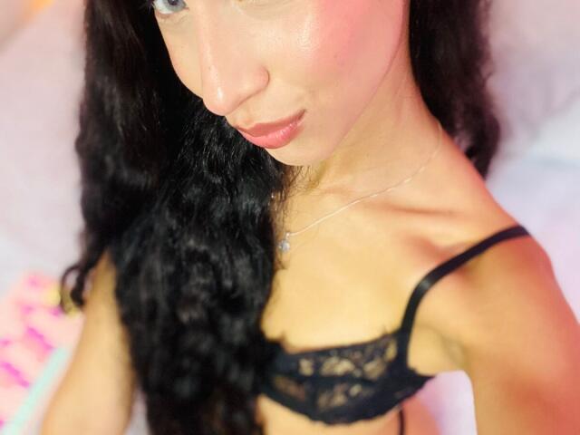 BeautifulShalom with black lace bra, wavy dark hair, pink blush, and subtle smile, showing her small tits and slim neck, wearing a delicate necklace.
