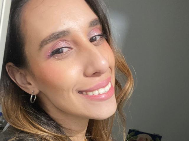 BellaBrownX's face, smiling with pink eyeshadow, brown hair with blonde highlights, hoop earrings, and pink lipstick, showing white teeth, with light skin and a slightly shiny forehead.