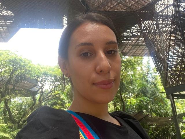 BellaBrownX has a cute face with light brown skin, dark hair, small nose, and subtle lips. She's wearing a black top with colorful straps, looking at the camera.