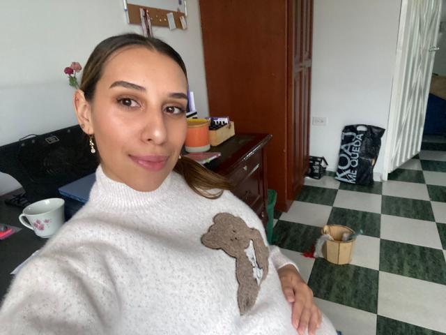 BellaBrownX with brown hair in a ponytail, wearing a white sweater with a brown tree design, smiling at the camera, standing in a checkered floor room.