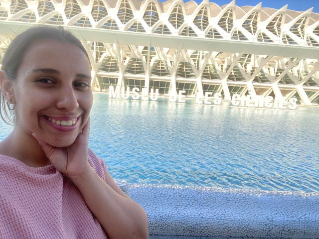 BellaBrownX with tan skin, brown eyes, and pink sweater, smiling, hands on cheeks, blue water pool in background, white geometric roof structure above, sunny day, relaxed.