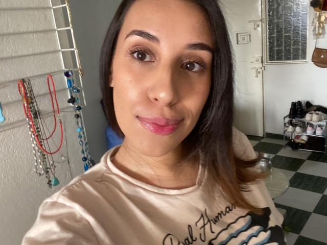BellaBrownX with shiny pink lips, brown eyes, and dark straight hair, wearing a beige shirt with a black graphic. Her face is slightly glossy. She's smiling at the camera.