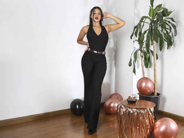 NaomiGray tem a pele morena, cabelo preto, e veste um top preto de alças finas com calças de cintura alta, mostrando suas curvas e seios pequenos. Ela está ao lado de uma planta e balões..