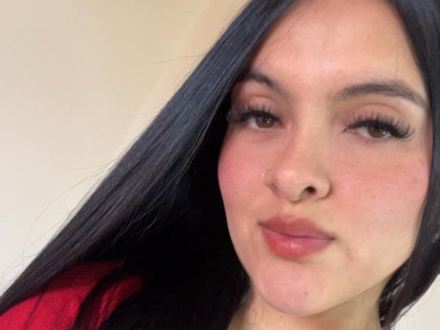 NicoleLie a de longs cheveux noirs, des yeux marron et des lèvres pleines roses, avec un petit anneau au nez. Elle porte un haut rouge, avec un sourire discret et un regard séduisant..