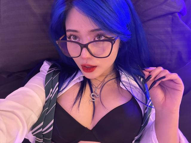 AmeliaXWalker with blue hair, glasses, and red lipstick, wearing a green striped tie, white shirt, black bra, and necklace, lying down, showing her tits.