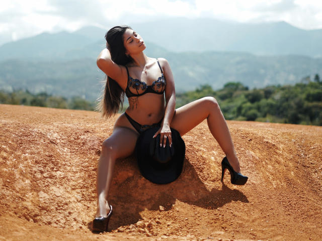 AnaWillsson sitting on a dirt mound, legs spread, wearing black lace bra and panties, holding a black hat, black high heels, long brown hair, tan skin, mountains in background.