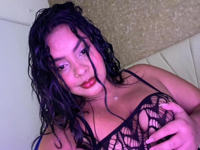 AmarantaCarey with wet black hair, pinkish skin, and red lipstick, wearing a black mesh top, holding her large tits with a slight smile, in a pink-lit room.
