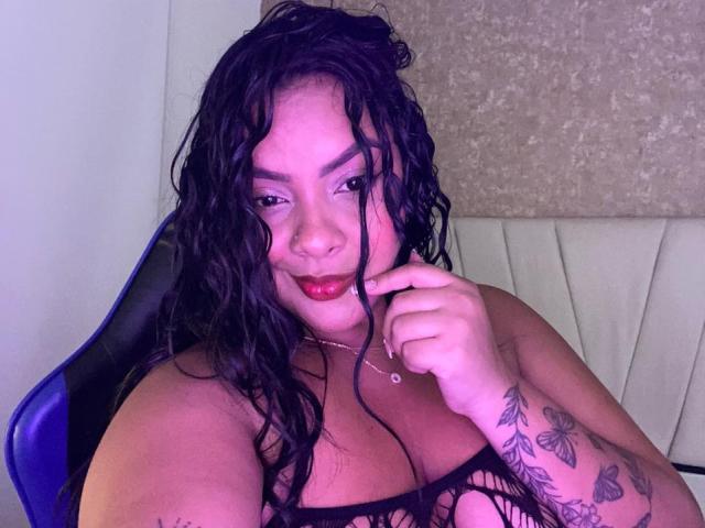 AmarantaCarey with wet black hair, red lipstick, and tattoos on her right arm, sitting in a blue chair, touching her lip with her finger, wearing a black top.