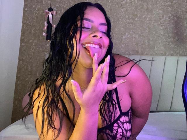 AmarantaCarey with wet black hair, smiling, tongue out, hand near mouth, wearing a black netted top, exposing large boobs, purple and pink lighting, in a bedroom.