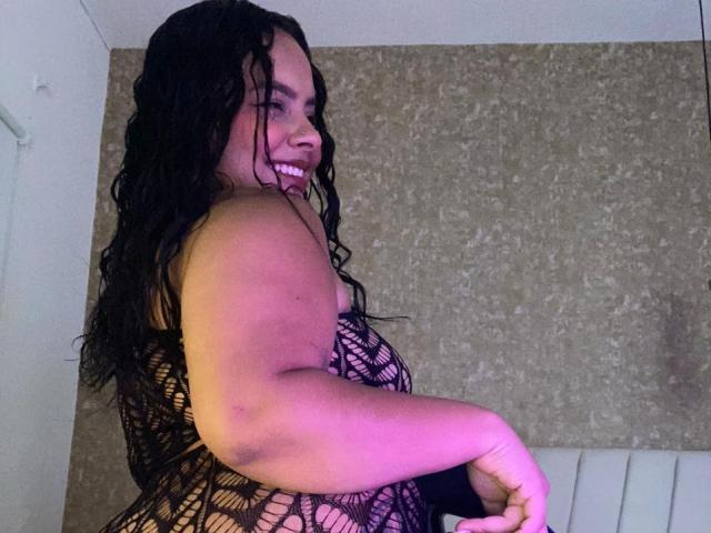 AmarantaCarey with wet black hair, smiling, wearing a black netted lingerie top, showing her curvy, tan-skinned body and small to medium tits, in a slightly turned pose.