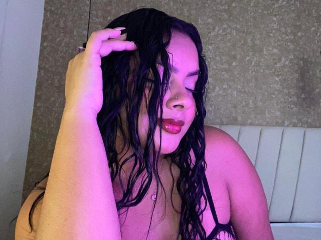 AmarantaCarey with wet black hair, pink-tinged skin, and glossy red lips, holding her hair, showing her bare shoulders and small tits, in a softly lit, purple-hued setting.