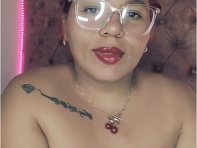 Cristina Mendez, met een bril, rode lippenstift en een gouden ketting met een rode kers, onthuld, met een tatoeage zichtbaar op haar rechter schouder, lachend. Haar borsten zijn zichtbaar, ze heeft lichtbruine huid..