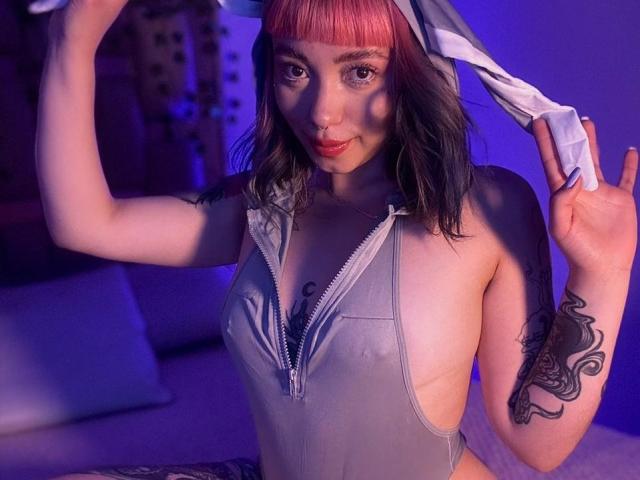 GigiVolkova with pink and black hair, holding a white headscarf, wearing a zip-up grey one-piece, showing a tattoo on her right arm, and a nose ring.