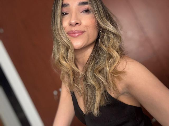 AdharaCooperr with wavy blonde hair, pink eyeshadow, and subtle pink lipstick, wearing a black dress, smiling slightly, showing her boobs and a cute face.