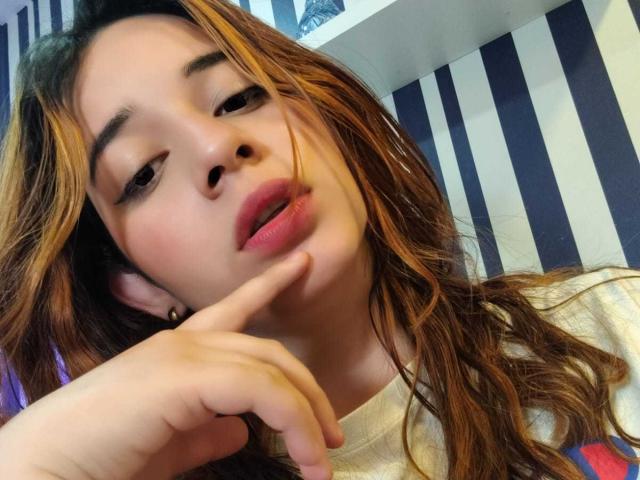 DaniVelasquez with her finger on her lips, pink lips slightly parted, brown wavy hair with blonde highlights, looking seductively at the camera. She's wearing a white shirt.