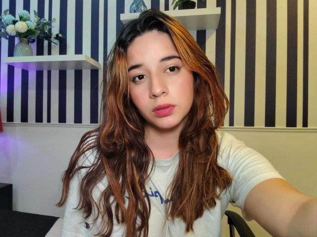 DaniVelasquez with wavy brown and blonde hair, pink lips, and light skin, wearing a white shirt, in a selfie, holding a phone, sitting in a chair.