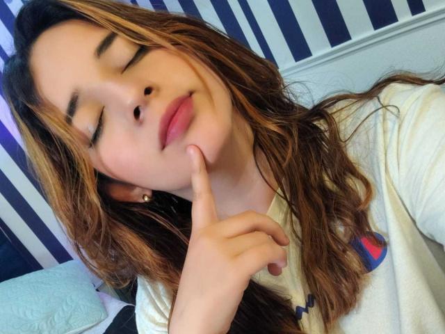DaniVelasquez has wavy brown hair, closed eyes, and a subtle smile. She's touching her cheek with her finger, wearing a white shirt with a colorful logo.
