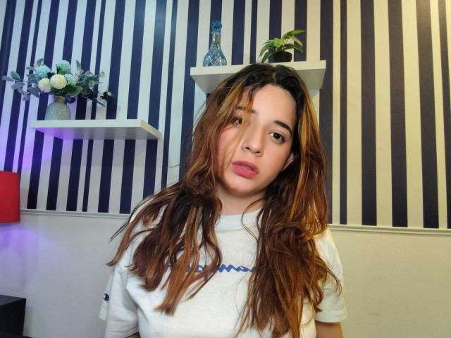 DaniVelasquez with wavy brown hair, wearing a white shirt, has a neutral expression, standing in a room with striped black and white wallpaper. She has small tits.