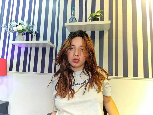 DaniVelasquez with wavy brown hair, pink lips, white t-shirt, standing in front of striped wall, shelves with vase and plant, looking at camera with slightly pouty expression.