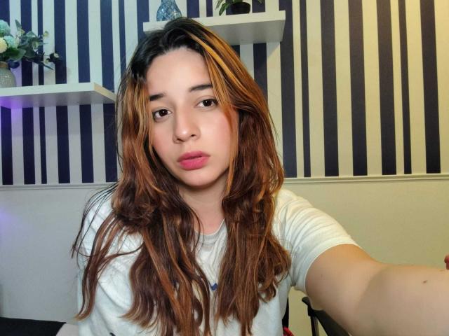 DaniVelasquez with long brown wavy hair, pink pout, white t-shirt, holding out phone for selfie, striped wall background, white shelves with small plants.