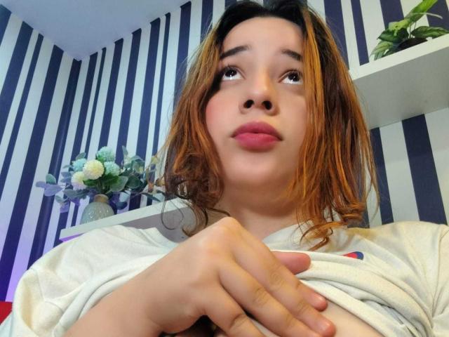DaniVelasquez with pink lips, brown hair, and white shirt, hands clasped, looking up with a soft expression, striped blue and white wall in background.