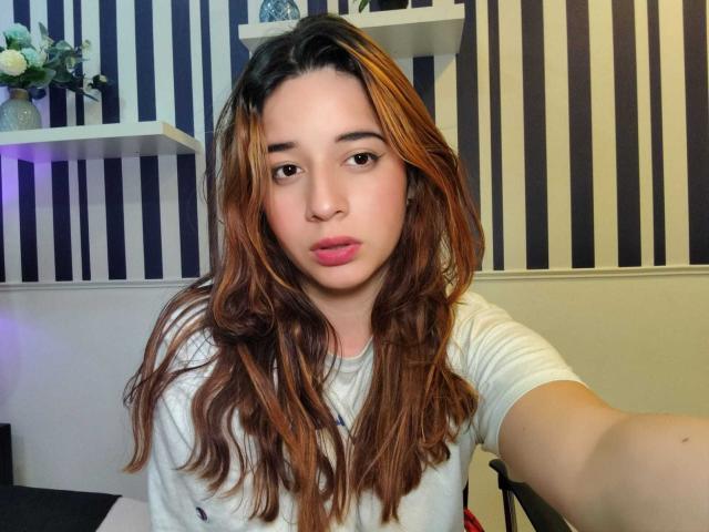 DaniVelasquez has long, wavy brown hair with blonde highlights, pink lips, and a white t-shirt, taking a selfie with a neutral expression, showing her cute face and boobs.