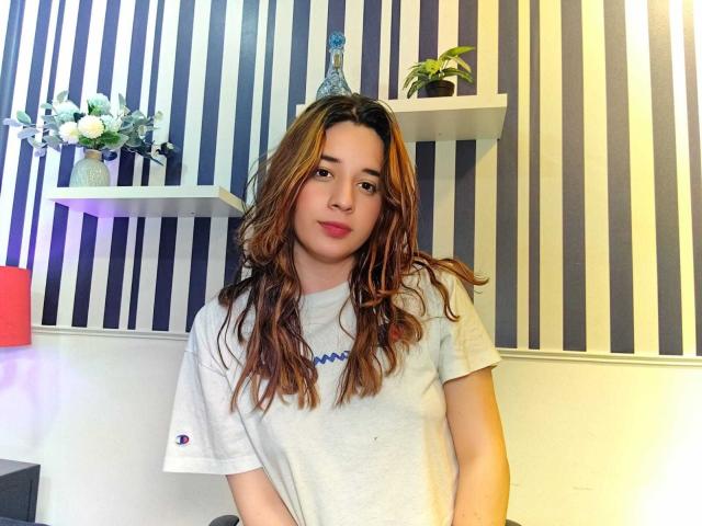 DaniVelasquez with wavy brown hair, wearing a gray t-shirt, standing in a room with blue and white striped walls, small shelves, and potted plants, smiling subtly.
