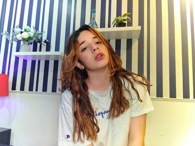 DaniVelasquez with long, wavy brown hair, pink lips, and white tee, leaning forward, looking at camera, showing a cute, relaxed expression, in a striped room.