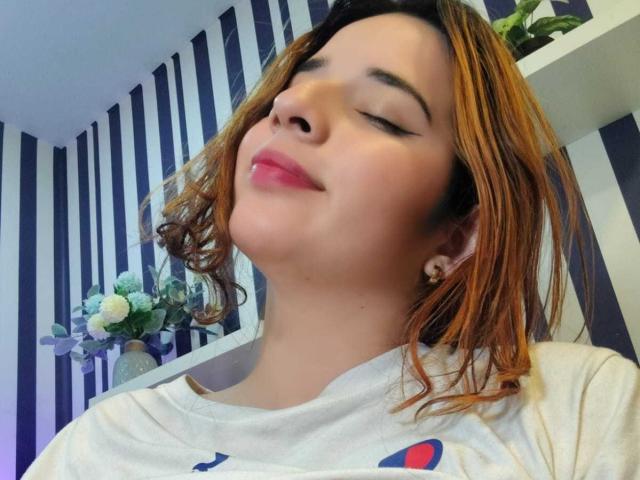 DaniVelasquez has her eyes closed, smiling with pink lipstick, and short brown hair, wearing a white shirt with a blue and red logo, in a striped room.