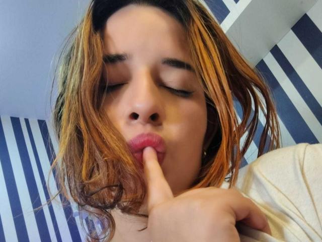 DaniVelasquez, with eyes closed, has messy brown hair, and is sucking on her index finger with pink, slightly parted lips. She's wearing a white shirt.