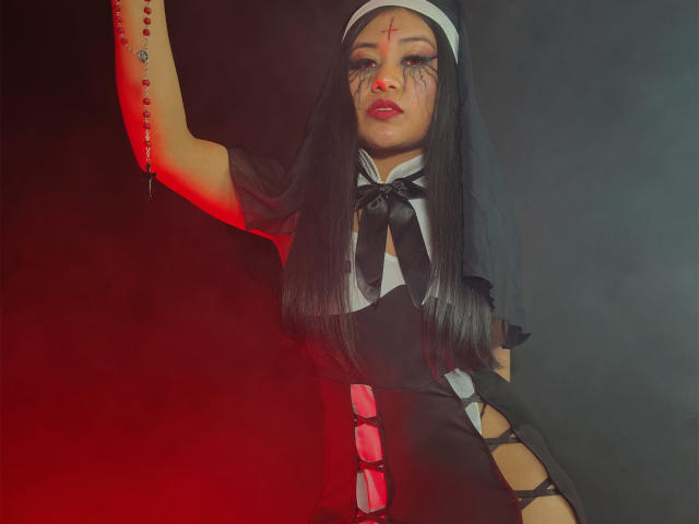 LiaDecker in a black nun outfit with long black hair, red lipstick, and a crucifix on her arm, raising her hand, showing a rosary, and revealing her tits.