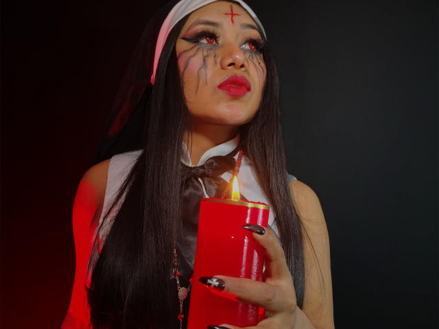 LiaDecker with long black hair, nun outfit, red eyes, black facial paint, holding red candle, red light, white headcover, black nails, red lipstick, looking up.