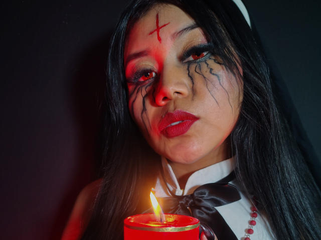 LiaDecker with dark makeup, red lipstick, and black cracks on her face, holding a lit red candle, wearing a white nun outfit with a black bow.