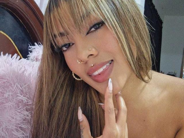 AmelieDuvall with long brown hair, bangs, and gold nose piercings, smiling, showing off pink nails, and wearing gold hoop earrings, in a selfie. Her tits are slightly visible.