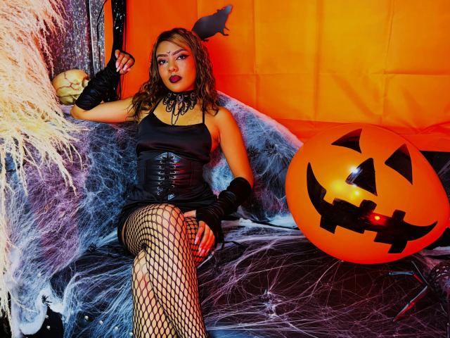 AmelieDuvall with curly brown hair, black corset, fishnet stockings, black gloves, and a black choker, sitting next to a glowing pumpkin, showing her tits and ass.