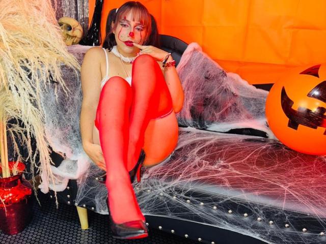 MilaWilde with red thigh-highs, white lingerie, and black heels, sitting on a spider-webbed couch, her ass raised, wearing a pearl necklace, next to a jack-o'-lantern.