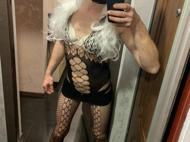 MilankAss with long white hair, black fishnet bodysuit, exposed breasts in white bra, and black mini skirt, taking a mirror selfie, showing off their taut, toned body.