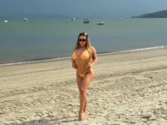 AgostinaNunier with big tits and a small orange bikini, standing on a sandy beach, wearing sunglasses, with long blonde hair and a curvy, sexy, attractive body.