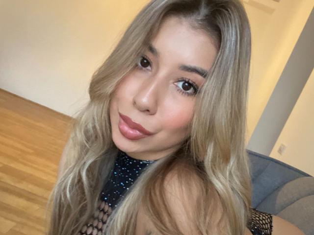 AgostinaNunier with blonde hair, pouty pink lips, and dark eyes, wearing a black mesh top, showing her boobs, in a selfie with a playful, slightly tongue-out expression.