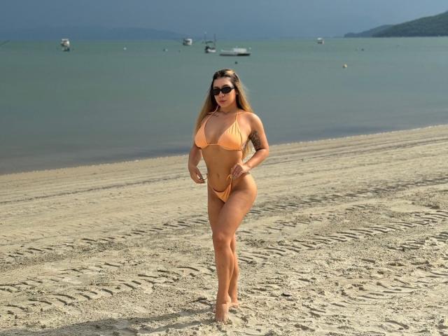 AgostinaNunier with big ass and big tits in an orange bikini, standing on a sandy beach, wearing black sunglasses, and walking towards the camera.