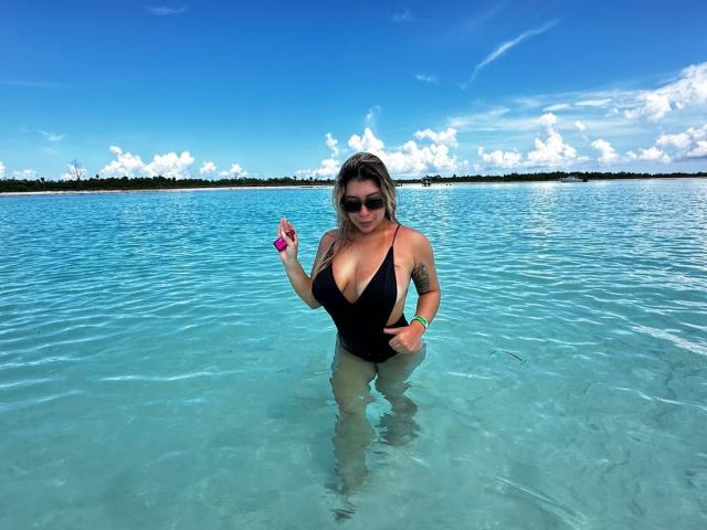 AgostinaNunier with big tits in a black one-piece swimsuit, wading in clear blue water, holding a pink object, wearing sunglasses, with a tattoo on her right arm.