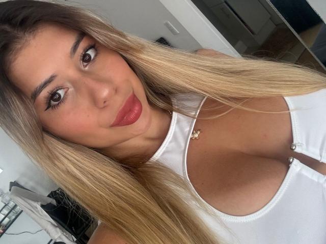 AgostinaNunier has long blonde hair, red lipstick, and a white zip-up top showing her large tits. She's looking at the camera with a slight smile.