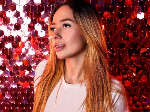 LinaMillia with long, straight, blonde-to-brown ombre hair, wearing a white top, has shiny pink lips and a slight smile, standing against a red, shiny, hexagonal-patterned background.