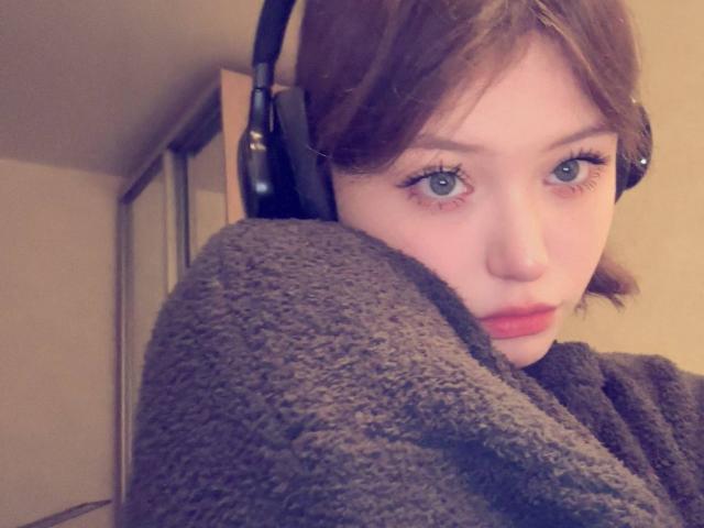 NikolKitt has blue eyes, brown hair, and is wearing black headphones with a gray textured sweater, looking serious with slightly parted red lips.