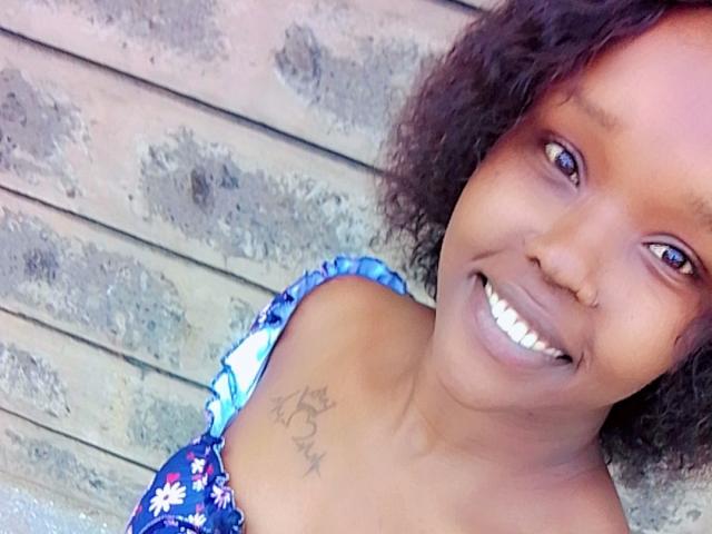 BustyNeptune has a cute smile with white teeth, brown curly hair, and a tattoo of stars on her shoulder. She's wearing a blue floral dress, showing her tits.