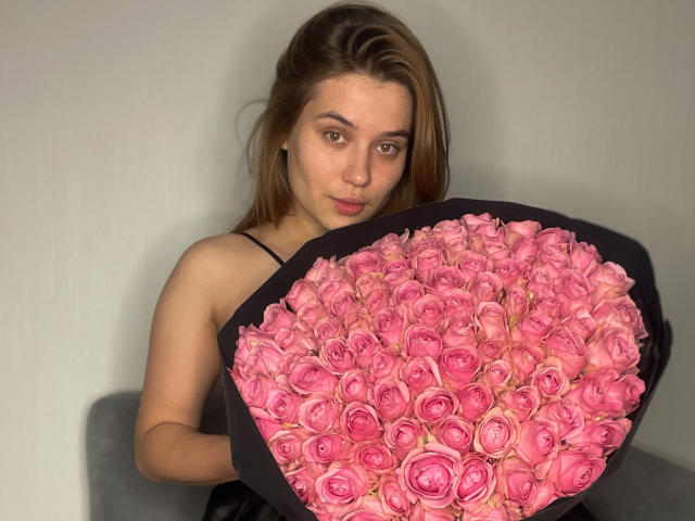 LioraEm with pink roses, holding a big black-wrapped bouquet, wearing a black spaghetti strap top, brown eyes, light skin, and brown hair, looking at the camera.