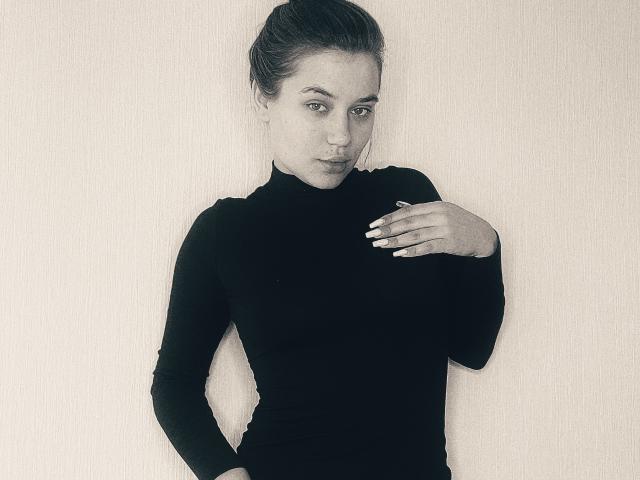 LioraEm with her hand on her chest, wearing a tight black turtleneck, her ass slightly visible, standing against a plain background, looking directly at the camera.