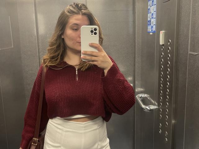 LioraEm with medium tits, wearing a red zip-up sweater and white high-waisted pants, taking a mirror selfie in an elevator, holding a white iPhone.